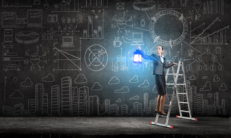Young businesswoman in darkness with lantern in hand standing on ladderの写真素材
