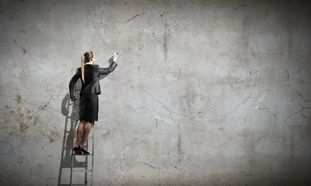 Rear view of businesswoman standing on ladder and drawing on wallの写真素材