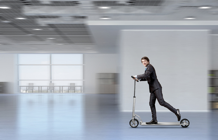Young cheeful businessman riding scooter in office. Mixed mediaの写真素材