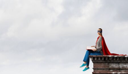 Young woman in mask and red cape sitting on building roofの写真素材