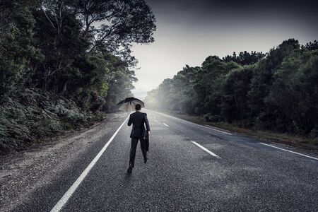 Back view of businessman with umbrella and suitcase walking on road . Mixed mediaの写真素材