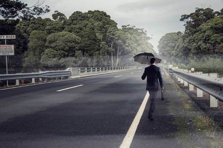 Back view of businessman with umbrella and suitcase walking on road . Mixed mediaの写真素材