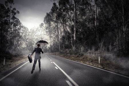 Back view of businessman with umbrella and suitcase walking on roadの写真素材