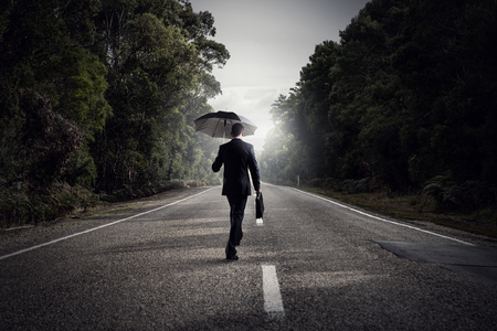 Back view of businessman with umbrella and suitcase walking on roadの写真素材