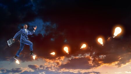 Black businessman running on hypothetical stairs made of burning lamps on a dark cloudy evening sky backgroundの写真素材