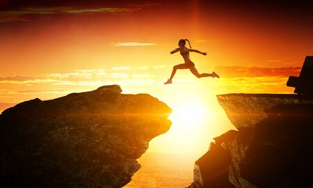 Woman jumping over mountain precipice in front of sunsetの写真素材