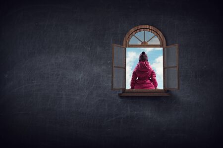 Kid girl in winter clothes sitting with back on window. Mixed mediaの写真素材
