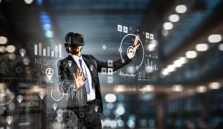 Young businessman wearing virtual helmet and working with media panel. Mixed media