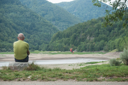Kruth - France - 25 August 2017 - man sitting on stone in border lake with paraglider on backgroundのeditorial素材