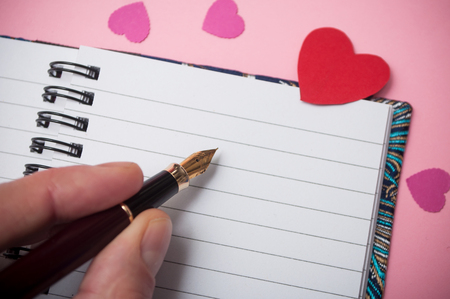 closeup of hand of man writing with vintage pen on spirales note book with paper hearts on pink backgroundの写真素材