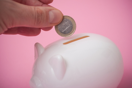 closeup of  hand of man putting euro coin in white piggy bank on pink backgroundの写真素材