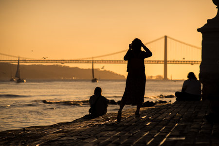 Lisbon - Portugal - 30 September 2020 - silhouette of tourist taking a picture with her smartphone of the famous 25th April suspension bridge on sunset backgroundのeditorial素材