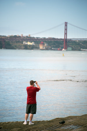 Lisbon - Portugal - 1 October 2020 - tourist taking a picture with his smartphone of the famous 25th April suspension bridge on blue sky backgroundのeditorial素材