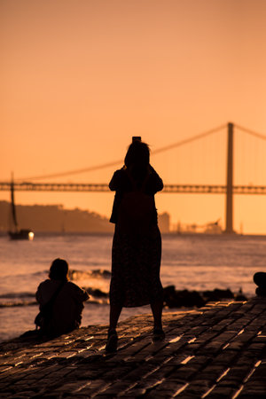 Lisbon - Portugal - 30 September 2020 - silhouette of tourist taking a picture with her smartphone of the famous 25th April suspension bridge on sunset backgroundのeditorial素材