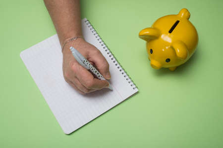 Closeup of hand of woman writing on  a note book with a yellow piggy bank on green background