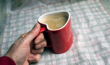 Closeup of red ceramic mug of coffee in shaped heart in hand on towel background