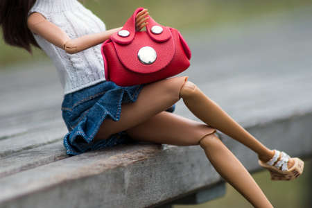 Closeup of legs of mannequin doll wearing a blue jeans skirt and red hand bag sitting on a wooden bench in a public gardenの写真素材