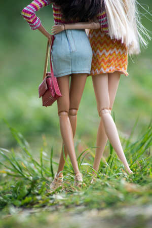 Closeup of blond and brunette barbie dolls legs wearing orange summer dress and blue jeans mini skirt  walking in a public gardenの写真素材