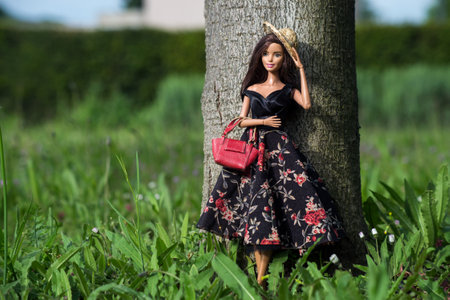 Mulhouse - France - 29 May 2021 - Portrait of brunette barbie doll wearing a black summer dress,  red leather handbag and summer hat  standing against a tree trunk in a public gardenのeditorial素材