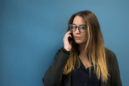 Portrait of young woman with eyeglasses using mobile phone on blue wall backgroundの写真素材