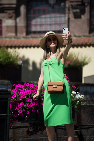 Colmar - France - 12 July 2022 - Portrait of pretty  woman wearing a green dress taking a photo with her smartphone in the streetのeditorial素材