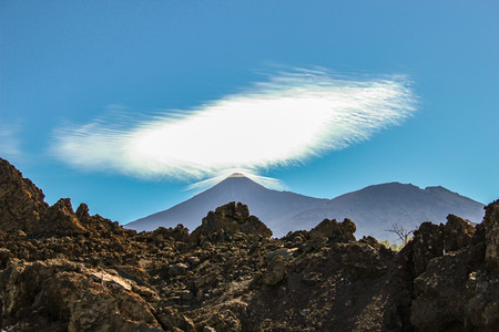 Valley covered by lava stones. Mountain Pico del Teide with white snow spots, partly covered by the clouds. Bright blue sky. Teide National Park, Tenerife, Canary Islands, Spain.の写真素材
