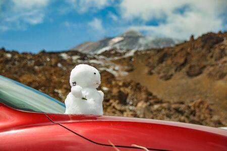 Base of volcano Teide. Snowman on on the hood of the red car. Peack of Teide with white snow spots, partly covered by the clouds. Bright blue sky. Teide National Park, Tenerife, Canary Islands, Spain.の写真素材