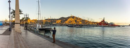 Cartagena, Spain - December 2, 2017: Yung man makes picture of promenade using smartphone. Panoramic view of Cartagena Murcia port marina sunset time in Mediterranean Spain.のeditorial素材