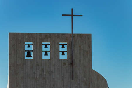 Cross on church over clear blue sky. Six bells of different sizes on the top. Adeje village,...