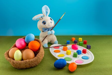Colored easter eggs in the nest of straw. Gray plush rabbit with a palette with a brush paints...