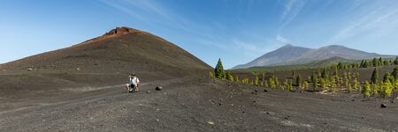 Super wide panoramic view of Volcano Arenas Negras and lava fields around. Bright blue sky and white clouds. Young traveler sits on a stone and rests. Teide volcano in the background. Tenerife.の写真素材
