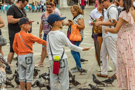 St. Mark Square, VENICE, ITALY - August 02, 2019: Warm sunny summer day. Numerous tourists are photographed with city pigeons. Children frolic and try to catch and play with birds.のeditorial素材