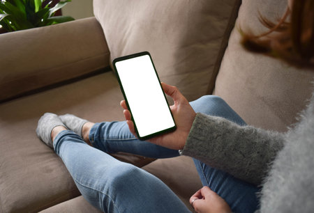 Image of a woman holding a mobile phone with a blank screen while sitting on the sofa with a feeling of relaxation.の写真素材