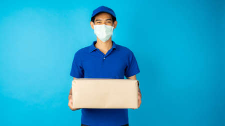 Asian delivery man wearing mask holding parcel box and show screen of phone on blue background in studioの写真素材