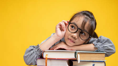 Thai Asian girl student fall down on stacked book, wearing glasses and looking camera in studio on orange or yellow backgroundの写真素材