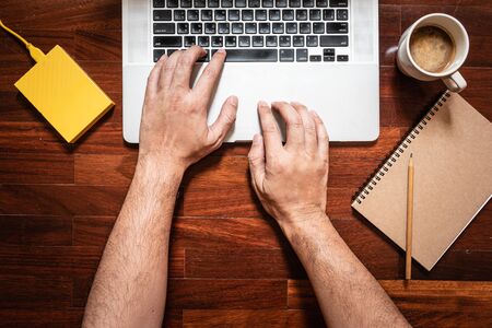 The man's hand typing the keyboard On the laptop computer there is a coffee mug, notebook, pencil...