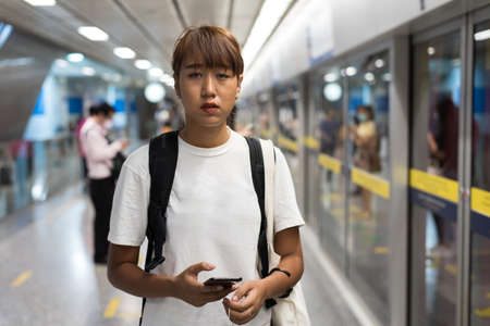 Asian woman look feel tired use smartphon standing waiting for subway, skytrain, carrying backpack, traveling to the city, social distancing, coronavirus, COVID19の写真素材