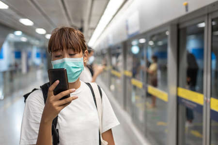Asian woman with surgical face mask feel tired use smartphone standing waiting for subway, skytrain,...