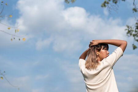 Young beautiful Asian woman wearing white shirt is in a cooldown position after exercising in the park. The background is part of the leaves of green trees. Blue sky And beautiful big clouds, view from the backの写真素材