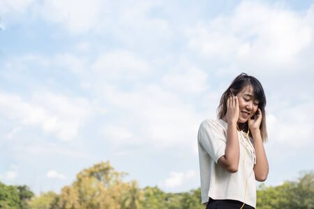 Beautiful Asian women wearing white shirts. Put on yellow headphones for listening to music, smile, and close her eyes feel happy in the garden. The background is part of the leaves of the green tree. Beautiful blue sky and white cloudsの写真素材