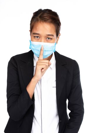 Asian businesswoman wearing surgical face mask in formal black suit jacket, looking at camera and place the finger on the mouth, studio lighting isolated on white background, coronavirus, COVID-19 conceptの写真素材