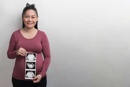 Smiling Asian pregnant woman holding ultrasound picture of her baby, look at the camera, isolated on grey backgroundの写真素材