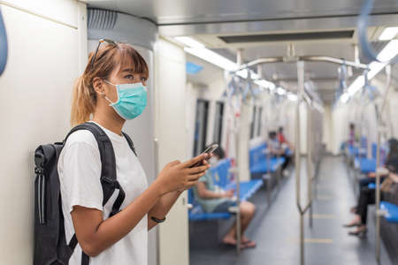 Side view of young Asian backpacker woman wear face mask holding smartphone travel to the city by...