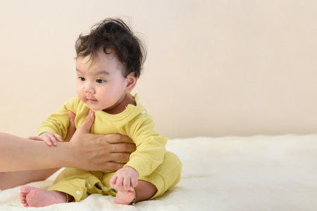 A cute little Asian baby in a yellow dress is lying on the white bed looking at the camera making a...