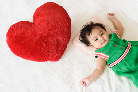 A cute little baby girl wear green dress lying beside a red heart shaped pillow on the bedの写真素材