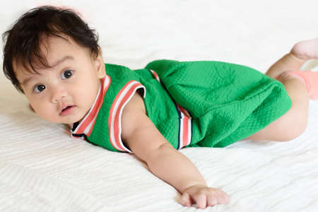A cute little baby girl wear green dress lying beside a red heart shaped pillow on the bedの写真素材