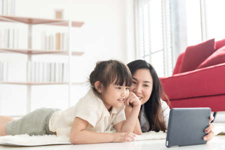 Happiness Asian young mother and little cute daughter lying on bed in the house using labtop for entertain, family joyful together in the house on holidayの写真素材