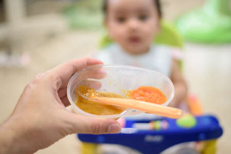 Hand holding a baby food bowl on a little baby's blurred background, focus on the food bowlの写真素材