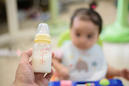 Hand holding a baby bottle of milk over blurred background of little baby, focus on bottleの写真素材