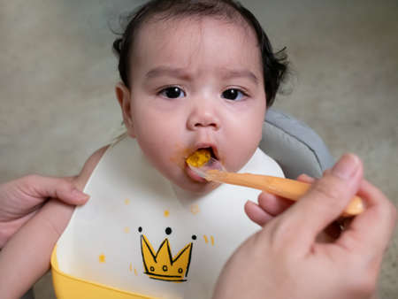 Mom feeding food her little Asian baby girl with a spoon at home